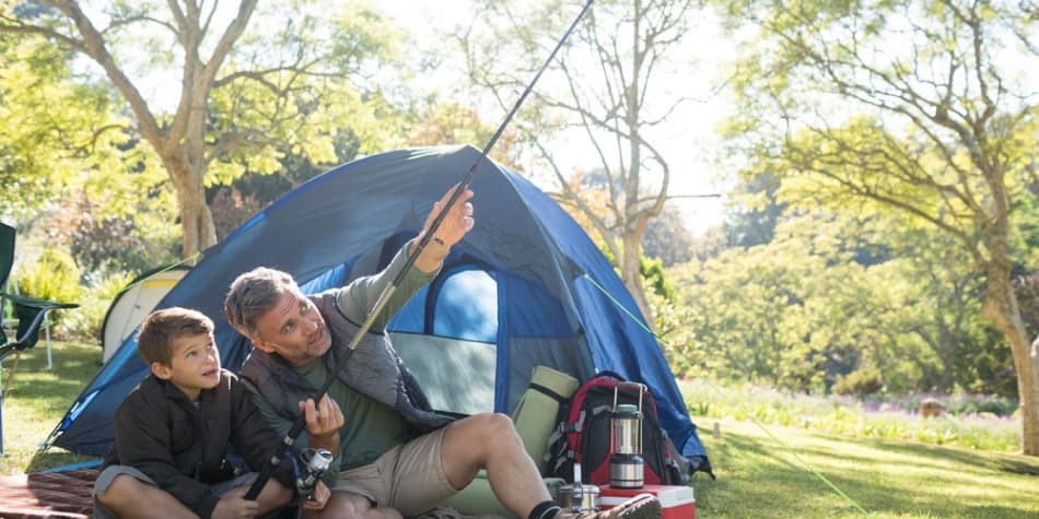 A father and son adjust a fishing pole outside of a tent while camping
outside.